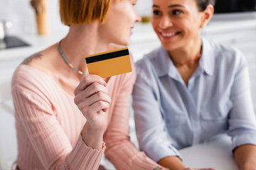 selective focus of credit card in hand of lesbian woman looking at cheerful african american girlfriend, blurred background