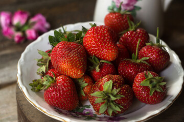 Ripe strawberries in a white old plate, near a rose. Brown wooden background