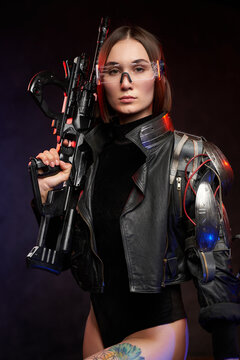 Futuristic portrait of a attractive female mercenary with fashionable style posing in dark background with rifle. Martial woman in cyberpunk style dressed in black clothing.