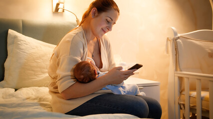 Smiling young mother feeding her newborn baby with breast milk working and browsing internet on smartphone at night in bedroom. Concept of healthy and natural baby nutrition. Health of mother and