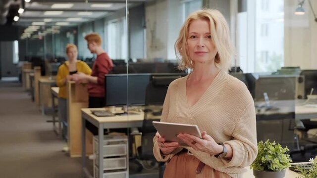Medium portrait of middle-aged caucasian female ceo of successful company standing with digital tablet in bright spacious office looking at camera