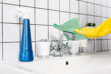 Cropped view of person in rubber glove cleaning faucet with rag near bottle of detergent on sink in...