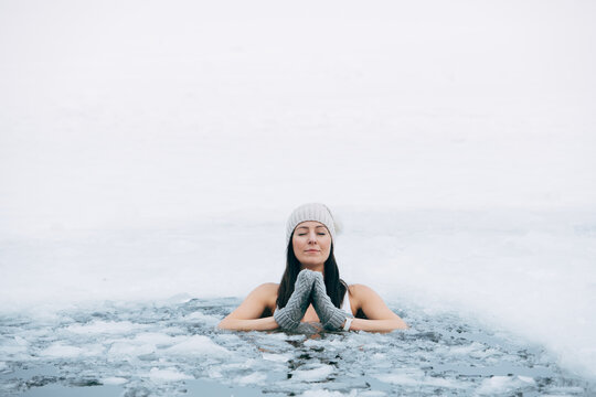 Winter swimming. Woman in frozen lake ice hole. Swimmers wellness in icy water. How to swim in cold water. Beautiful young female in zen meditation. Gray hat and gloves swimming clothes. Nature lake