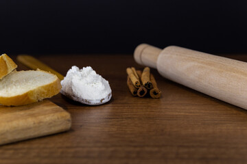 Fototapeta na wymiar Flour in a wooden spoon, sliced French baguette on a wooden board, rolling pin and cinnamon sticks on a wooden table. Top view with copy space. Kitchen baking tools with place for text, or logo.