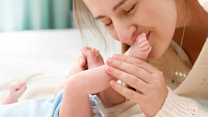 Closeup of young mother lying on bed and kissing little feet of her newborn baby son
