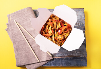 Noodles with chicken and vegetables in a box on yellow background and chopsticks.