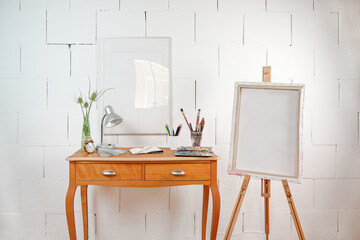 Tiny workplace for creative art work or hobby, crayons, sketchbook, watercolor and brushes on a wooden table and an easel with an empty picture frame, rough white painted wall, copy space