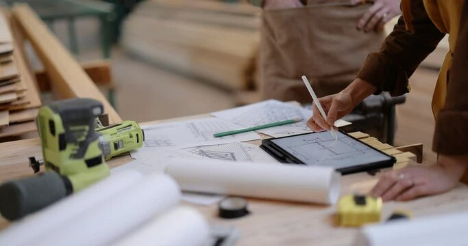 Craft persons designing some woodwork, drawing on a digital tablet at the workbench with tools and blueprints in the carpentry. Pan shot