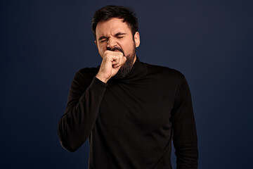 Handsome guy being bored talking with stranger about random staff, yawning cover opened mouth with fist, squinting tired, standing with fatigue look, dont like boring events, blue background