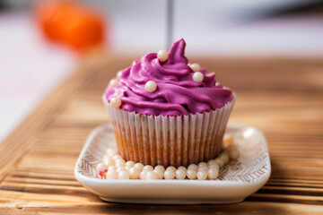 muffin with lilac color cream decorated with edible pearls on a saucer stands on a wooden background. Festive cake with cream. cream cheese, butter cream. Fresh bakery. Birthday dessert. small cake. 
