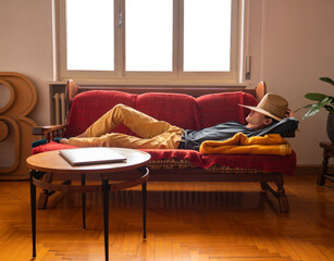 Young man wearing a hat sleeping on the sofa, lazy boy takes a nap during break to recharge his...