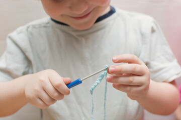 Happy child holds a crochet hook and blue thread in his hands, learns to knit. An interesting activity for a child, learning new skills. Horizontal photo.