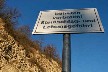 Sign before a yellow quarry saying: Do Not Enter! falling rocks and danger to life! ( germann: betreten verboten! steinschlag und lebensgefahr! ). Blue Sky in the Background.