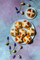 Homemade Easter traditional hot cross buns on ceramic plate with muscari flowers, quail eggs, textile over blue texture background. Flat lay, space