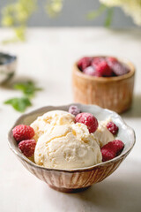Homemade caramel vanilla ice cream with frozen raspberries in ceramic bowl standing on white marble table with flowers behind.