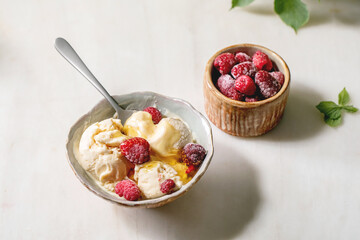 Homemade melted caramel vanilla ice cream with syrup and frozen raspberries in ceramic bowl standing on white marble table.