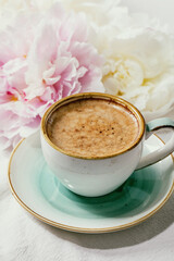 Cup of espresso coffee, pink and white peonies flowers with leaves over white cotton textile background.