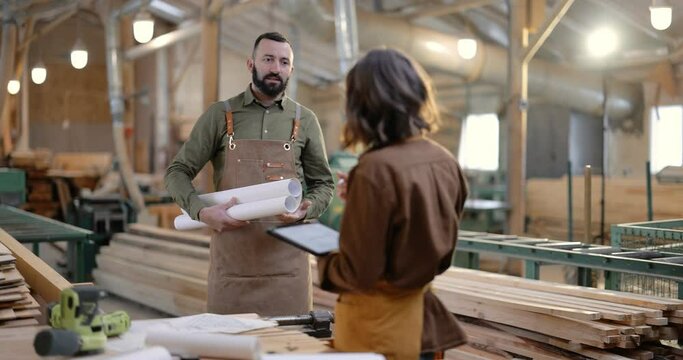 Carpentry workers discuss some working moments, standing with blueprints at the manufacturing. Teamwork, creative work, designing woodwork at the joinery