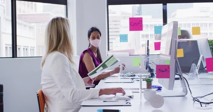 Diverse businesswomen wearing face masks using computers passing paperwork in office