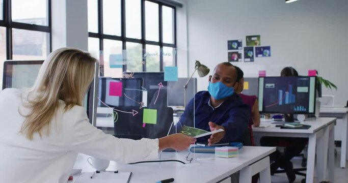 Diverse business colleagues wearing face masks using computers passing paperwork in office