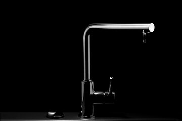 A modern metal kitchen water tap with a drop of water hanging down from it, isolated on the black background. Selective focus on the drop of water.