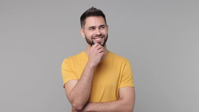 Smiling pensive bearded young man 20s years old in yellow casual basic t-shirt isolated on grey color background studio. People emotions lifestyle concept. Looking aside up put hand prop up on chin