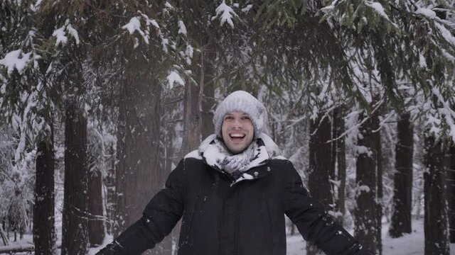 Medium shot, lot of snow is falling on face, hat and jacket of caucasian man from branches of fir tree in forest, avalanche of white snow comes down on man in winter, he laughs and smiles
