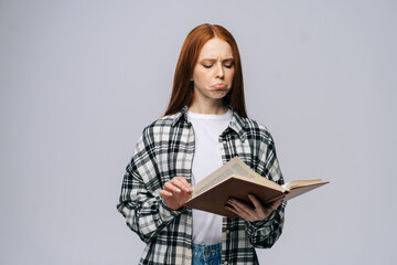 Obraz premium Confused young woman college student turning pages of book on gray isolated background. Pretty redhead lady model wearing casual clothes emotionally showing facial expressions in studio, copyspace.