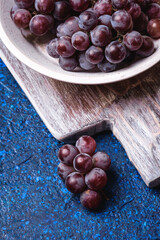 Obraz premium Fresh ripe grape berries in white wooden bowl and old cutting board on blue abstract background, angle view macro