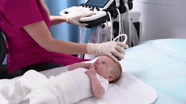 A female doctor conducts an ultrasound examination of the head and brain of a newborn baby using modern medical equipment in a clinic. Pediatrics and control of the development of the child's body.