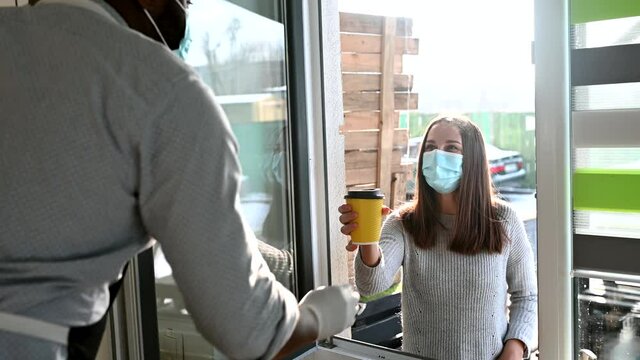 A male cafe worker gives a cup of takeaway coffee through a window to female customer wearing mask, small business works in conditions coronavirus pandemic