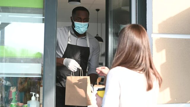 An african waiter wearing mask and gloves giving a paper cup of hot coffee to a female customer through an improvised take-away window for safety. Cafe works in take out due coronavirus pandemic