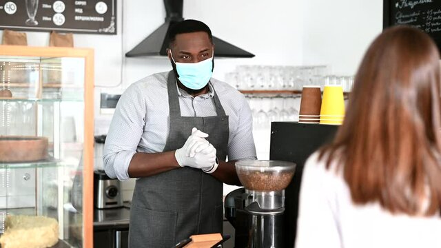 An African-American waiter wearing medical mask and gloves gives a package with takeaway food order to female customer, small business works in conditions coronavirus pandemic