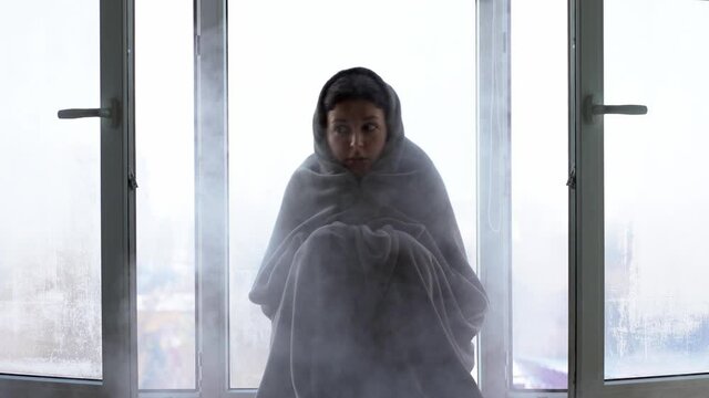 Cold winter weather. A woman is shaking from the cold sitting on the windowsill, wrapped in a blanket. Frosty air rushes in from the open window. Airing housing in cold winter