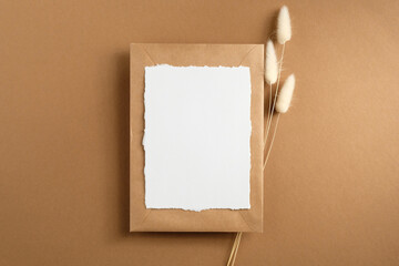 Empty paper card and kraft paper envelope with dried flowers on brown background. Flat lay, top view.