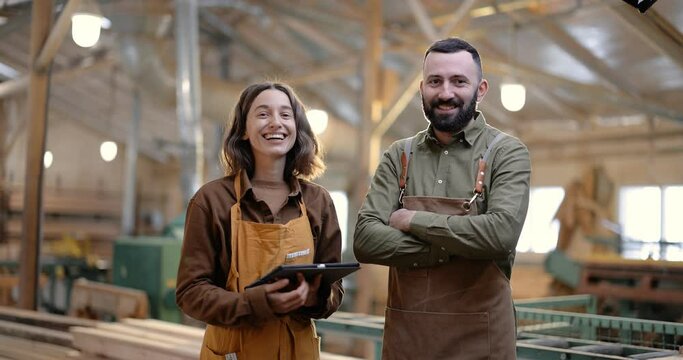 Portrait of two happy carpentry workers standing together in the joinery manufacturing. Designing woodwork or supervising production with a digital tablet