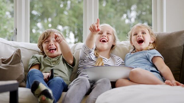 Three children sitting on sofa at home laughing and watching TV with popcorn together - shot in slow motion