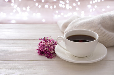A white cup of coffee, a bouquet and a sweater on the background of a burning garland. Side view with copy space. The concept of holiday backgrounds.