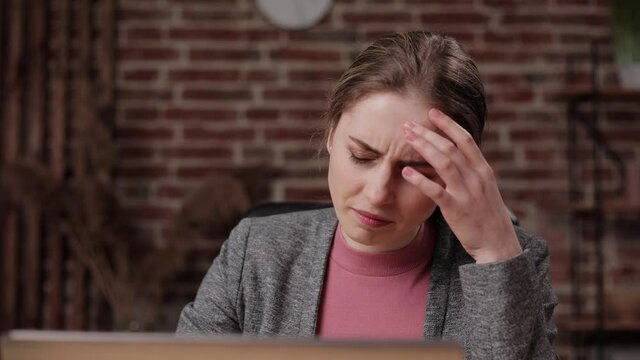 Brunette female with a headache take off her glasses. Woman working on computer getting bad headache massaging her head. Overworked business girl sitting in office workplace suffering from pain.