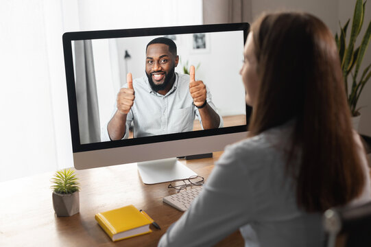 An African-American online coach, mentor, tutor on the laptop screen shows thumbs up. Back view a woman watching webinar, has video meeting with a biracial man