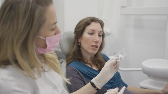 Professional orthodontist talks to patient about invisible retainer or braces. Female dental doctor or dentist explains and shows modern removable transparent plastic aligners for teeth alignment