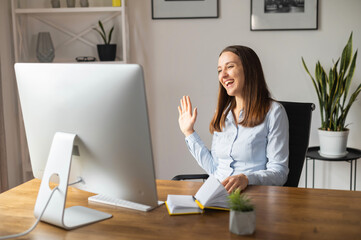 Cheerful female office employee in smart casual wear waving into webcam, talking online in virtual meeting sitting at the desk with PC in office