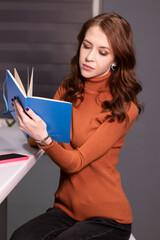 charming brunette woman in brown golf with blue notebook and phone in pink case sitting at table in gray room. work, education, study