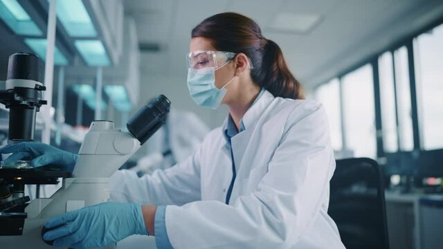 Female Scientist in Face Mask and Glasses Looking a Petri Dish with Genetically Modified Sample Chemicals Under a Microscope. Microbiologist Working in Modern Laboratory with Technological Equipment.