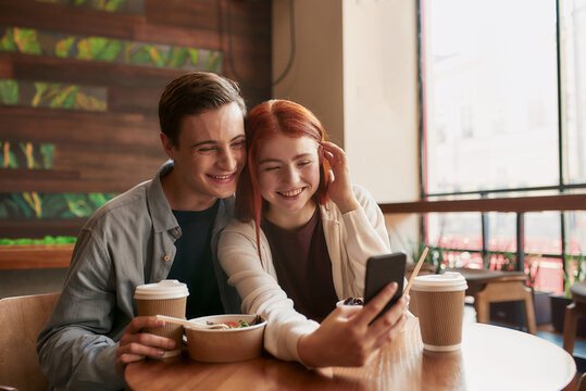 Happy teenage couple smiling at camera while taking selfies using smartphone, sitting in a cafe together on a daytime