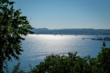 View of Novorossiysk and the port of Novorossiysk. Summer landscape on a sunny day. The sun is reflected in the water. Port cranes in the background