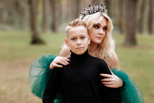 young beautiful blonde woman queen with young boy in black outfit. Princess mother walks with son. autumn green forest mystic. Vintage medieval shiny crown. Long evening green dress. magic fantasy