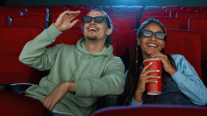 Portrait of cheerful young couple wearing 3d glasses smiling while watching a movie together in cinema auditorium and drinking soda