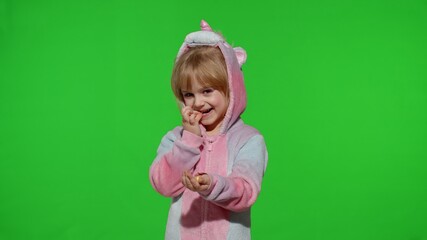 Portrait of a cute little child smiling, enjoying eating sweets candies dessert in unicorn costume on chroma key green background. Model kid girl animator in unicorn pajamas. Copy space
