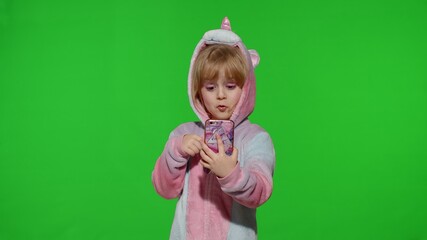 Excited little child girl in unicorn pajamas using, browzing on mobile cell phone, found out great big win news. Kid doing winner gesture isolated on chroma key background. Technology for children